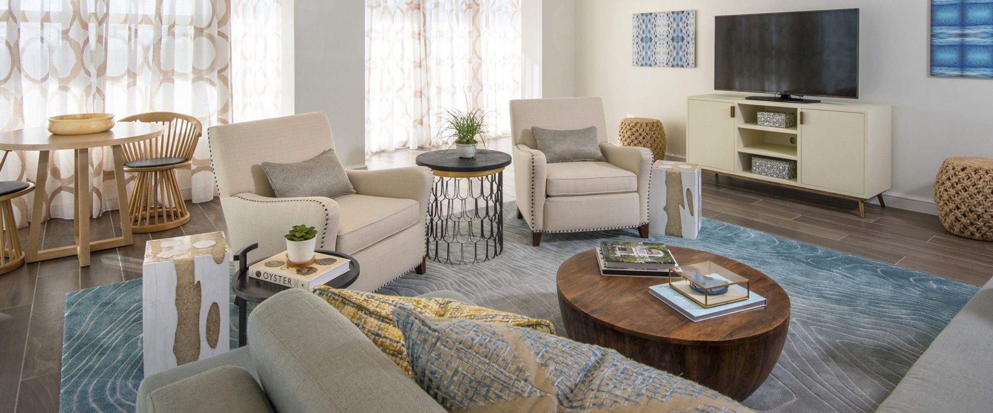 A modern living room with neutral-toned furniture, a round wooden coffee table, a flat-screen TV, patterned curtains, and various decorative items.