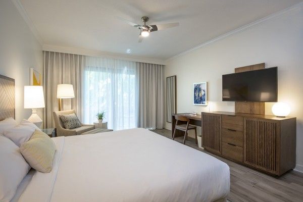 A modern hotel room with a large bed, chair, lamp, a dresser with a TV, desk, and a large window with curtains.