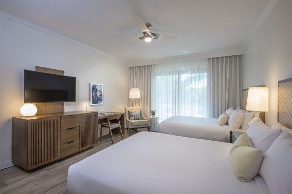 A hotel room with two beds, a wooden dresser with a TV, a desk and chair, floor lamp, and large window with curtains.