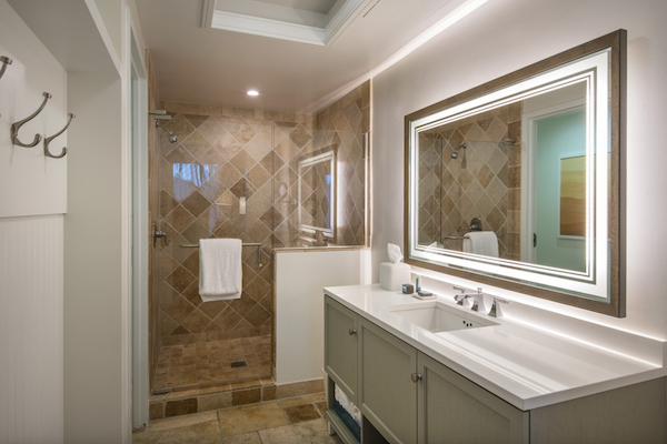 This image shows a modern bathroom with a tiled shower, a large rectangular mirror, a double sink vanity, and bright lighting.