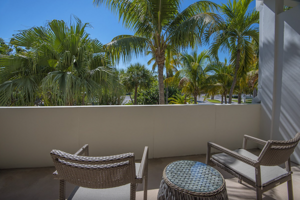 The image shows a balcony with two chairs and a small table, overlooking lush palm trees and a clear blue sky.