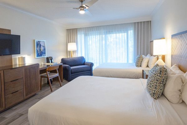 A hotel room with two beds, a desk, a TV, a sofa chair, lamps, and a large window with curtains. It looks bright and cozy.