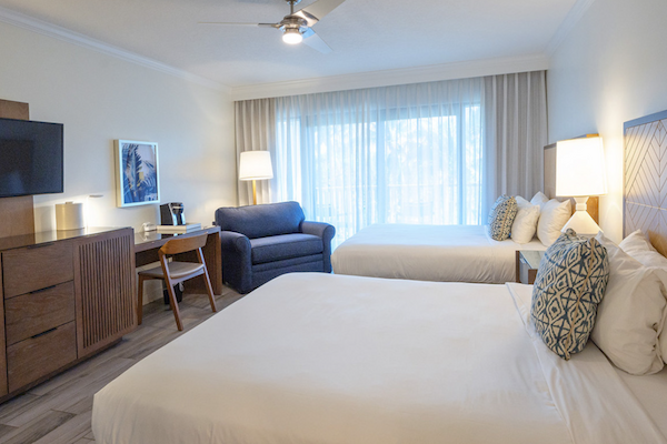 A hotel room with two beds, a TV, a desk and chair, a lounge chair, and large windows with sheer curtains, illuminated by lamps.