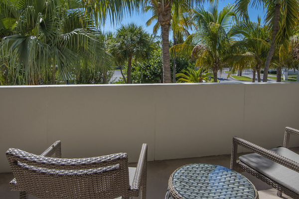 A balcony with two wicker chairs and a round table, overlooking lush palm trees in a sunny setting.