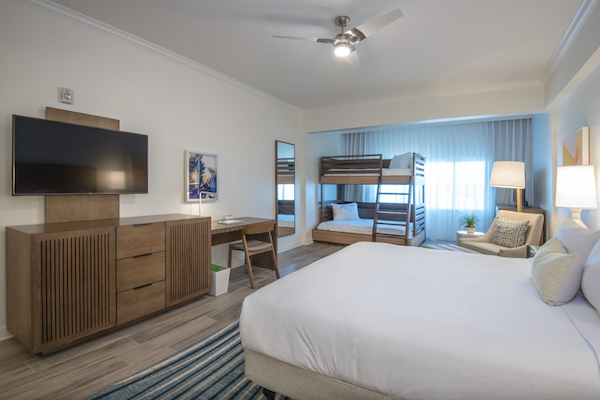 A hotel room with a bed, bunk beds, TV, desk, and lamps, featuring modern decor and natural lighting through the window.