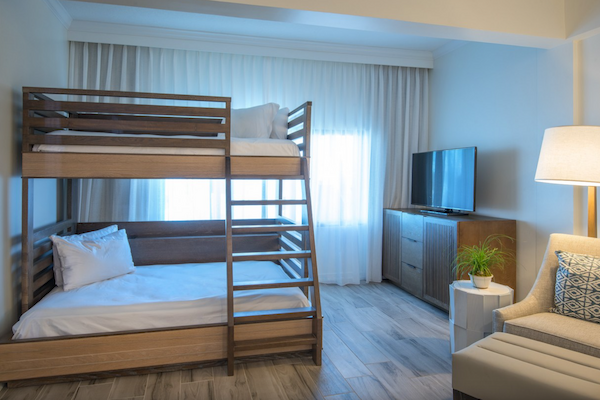 This image shows a room with a bunk bed, a TV on a cabinet, a lamp, and a chair with a cushion, all set against a backdrop of light curtains.