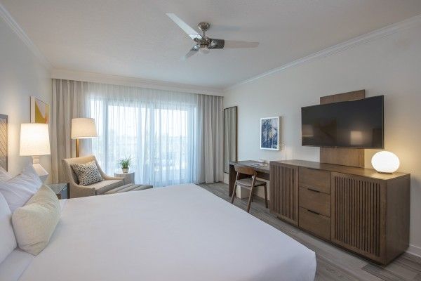 A modern hotel room with a large bed, a chair by the window, a TV on a dresser, a lamp, and a desk with a chair.