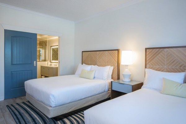 A clean, modern bedroom with two double beds, a nightstand with a lamp between them, and a blue door leading to a bathroom in the background.