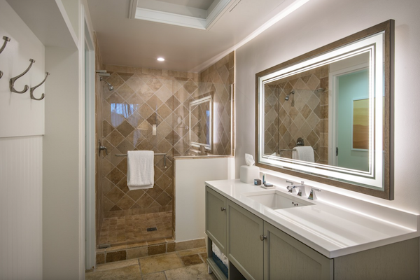 The image shows a modern bathroom with a large mirror, double sink, wall hooks, and a walk-in shower with a glass door and tiled walls, ending the sentence.