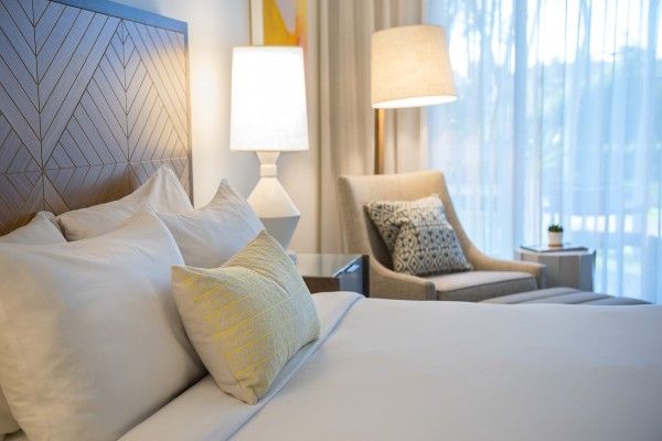 A neatly arranged bedroom with white bedding, two lamps, an armchair, a side table, and sheer curtains letting in natural light ending the sentence.