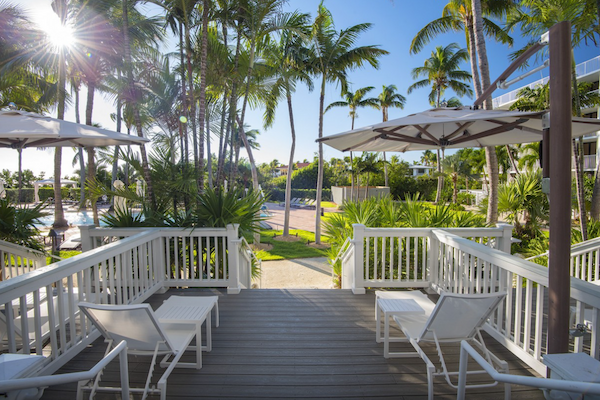 The image shows a tropical outdoor setting with palm trees, a bright sun, white lounge chairs, umbrellas, and a walkway leading to a pool area.