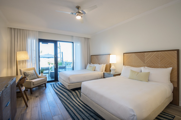 A hotel room with two beds, a desk, chair, lamps, and a window view of a balcony with outdoor furniture.