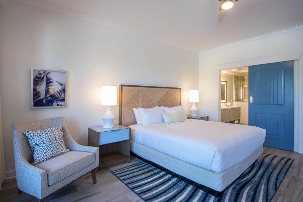 The image shows a modern hotel room with a bed, side tables, lamps, an armchair, decorative pillows, a painting, and a blue-striped rug.