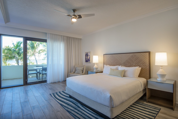 The image shows a bright, modern bedroom with a king-size bed, nightstands, lamps, and a seating area near large glass doors leading to a balcony.