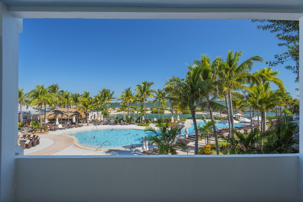 View from a balcony of a tropical resort featuring a pool, palm trees, sun loungers, cabanas, and a clear blue sky, creating a serene oasis.