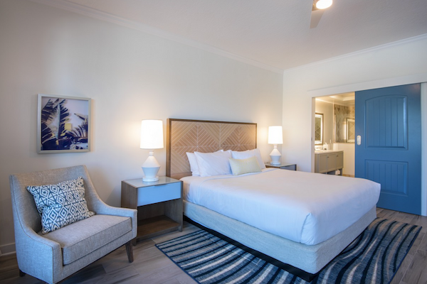 A cozy bedroom with a white bed, two bedside tables with lamps, an armchair, a striped rug, and a partially open blue door to a bathroom.