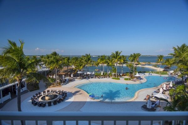 A resort featuring a large, curvy pool surrounded by palm trees and lounge chairs, with a beach and ocean view in the background.