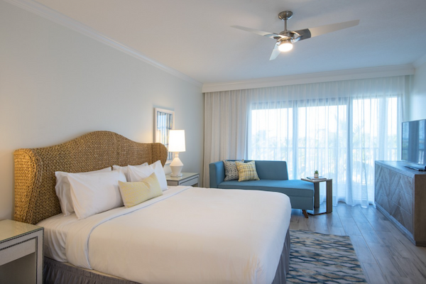 A cozy hotel room with a large bed, wicker headboard, bedside table, lamp, blue sofa, television on stand, ceiling fan, and large window.