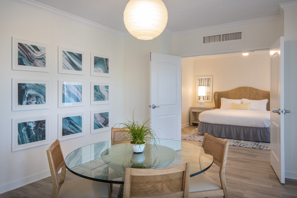 The image shows a cozy modern bedroom and dining area with minimalist decor, including a round table, chairs, potted plant, and framed art.