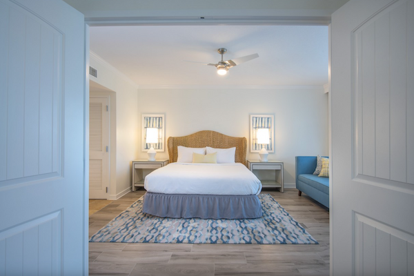 The image shows a neatly arranged bedroom with double doors, a large bed, bedside tables with lamps, a blue couch, and a patterned rug on wooden floors.