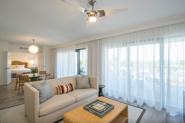 A modern living room with a sofa, colorful pillows, coffee table, dining area, and large windows with sheer curtains.