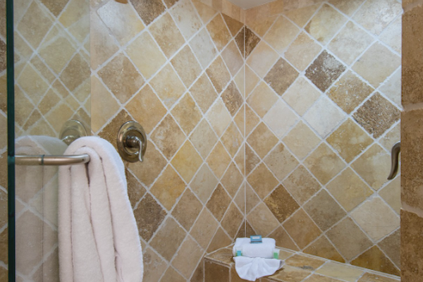 The image shows a tiled shower with a bench, two showerheads, a towel hanging on a rail, and a folded towel on the bench under a light.