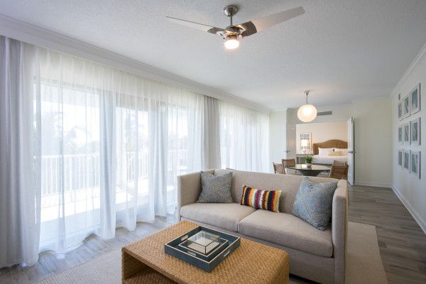 A modern living room with a beige sofa, wicker coffee table, and sheer white curtains, transitioning into a dining area with a round table.