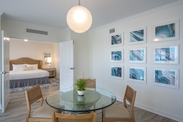 A modern room with a glass table, four chairs, and a small plant. Wall art decorates the space; the bed is visible through an open door.