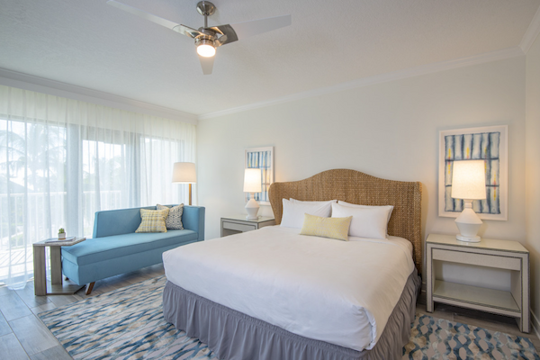 A bright, modern bedroom with a large bed, two side tables with lamps, a blue sofa, a ceiling fan, and sheer curtains letting in natural light.