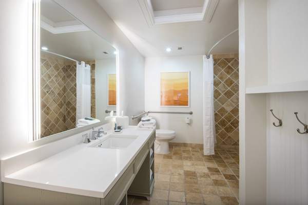 A clean, spacious bathroom with a double sink, large mirror, shower curtain, and neutral-toned tiles. There's wall art and hooks visible.