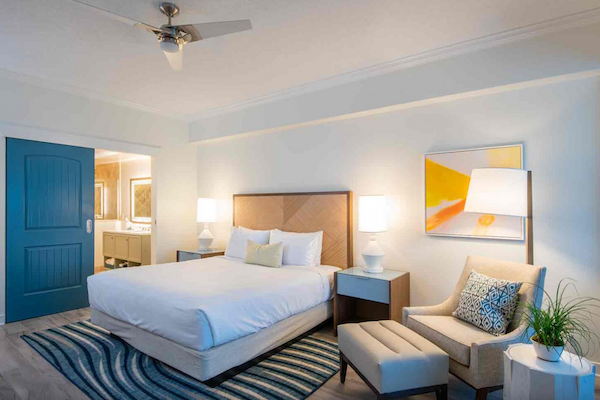 A well-lit hotel room with a bed, two nightstands, a chair, artwork, and a ceiling fan, featuring a modern design and cozy atmosphere.