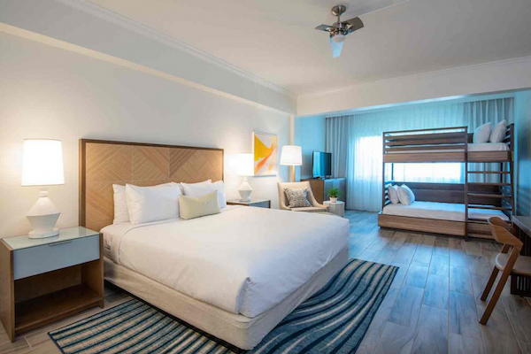 A spacious hotel room with a king-sized bed, bunk beds, desk, table lamp, and modern decor. The floor is wooden with a striped rug.