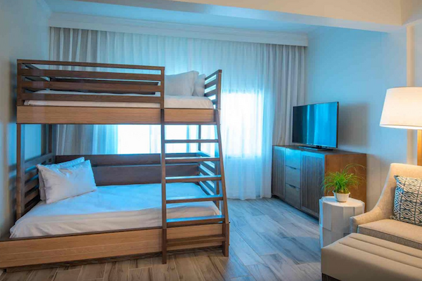 A room with bunk beds, a TV on a dresser, a cozy chair, and a potted plant, all lit by a floor lamp and natural light through the curtains.