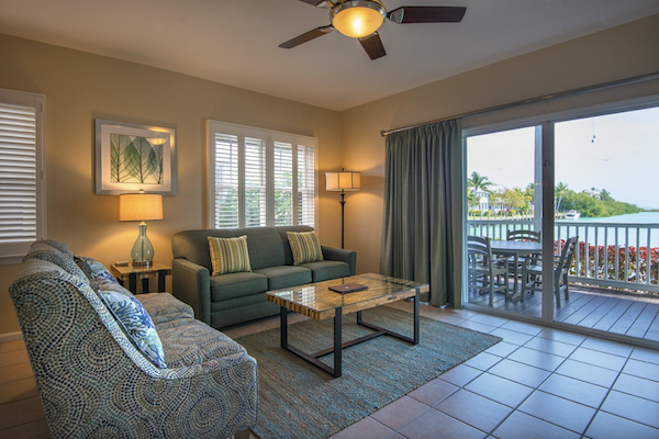 A cozy living room with a sofa, armchairs, coffee table, and a ceiling fan. There's a sliding glass door leading to a balcony with a scenic ocean view.