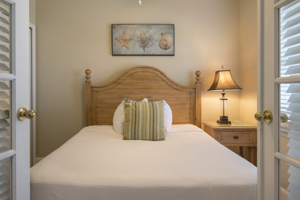 A cozy bedroom with a neatly made bed, flanked by nightstands and lamps, a ceiling fan, and decor above the bed, viewed through double glass doors.