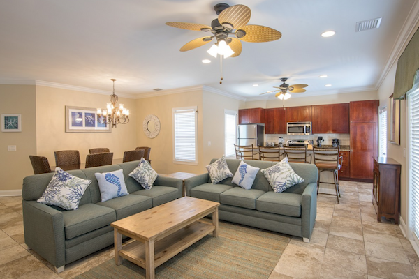 This image shows a modern open living area with two grey sofas, a wooden coffee table, a kitchen, and a dining area with a table and chairs.