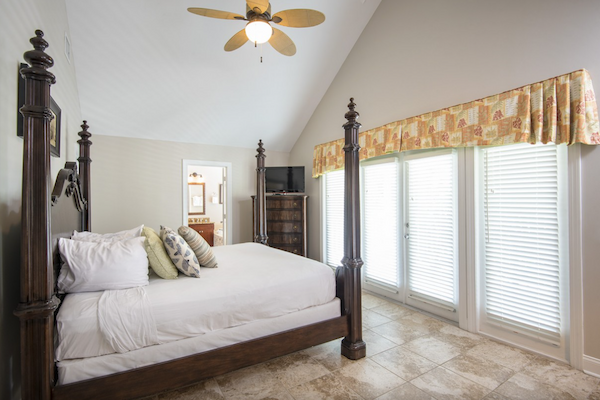 A stylish bedroom with a four-poster bed, ceiling fan, TV, and large windows with blinds and a patterned valance.