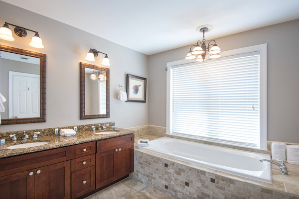 The image shows a modern bathroom with a double vanity, large mirrors, a bathtub, tiled walls, and a window with blinds.