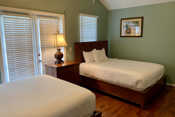 A bedroom with two beds, a nightstand with a lamp, a painting on the wall, windows with blinds partially open, and wood flooring.