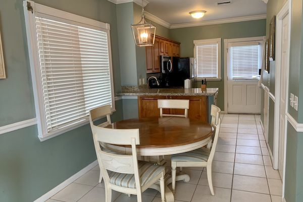 The image shows a kitchen with light green walls, white blinds, a round wooden dining table with four chairs, an island, and wooden cabinets.