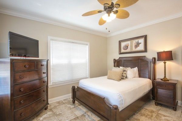 A bedroom with a wooden bed, dresser with TV, nightstand with lamp, ceiling fan, window with blinds, sea-themed wall art, and tile flooring.