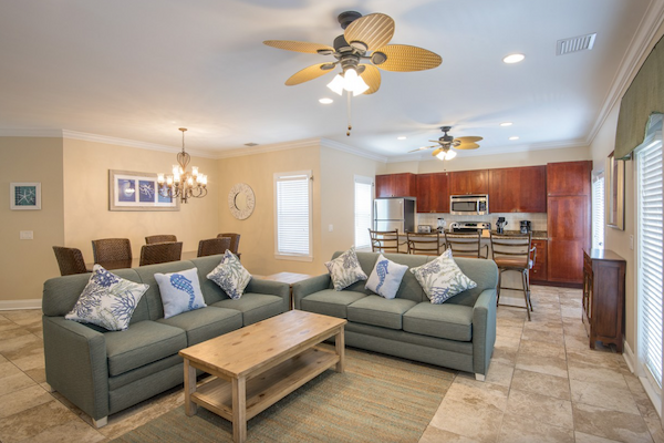 A modern open-plan living area with sofas, a kitchen, and a dining table. The space is bright and well-lit with ceiling fans.