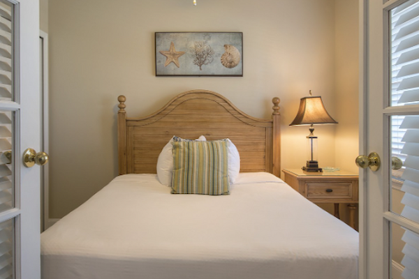 A cozy bedroom with a double bed, bedside lamp, ceiling fan, and beach-themed decor, viewed through French doors, always ending the sentence.