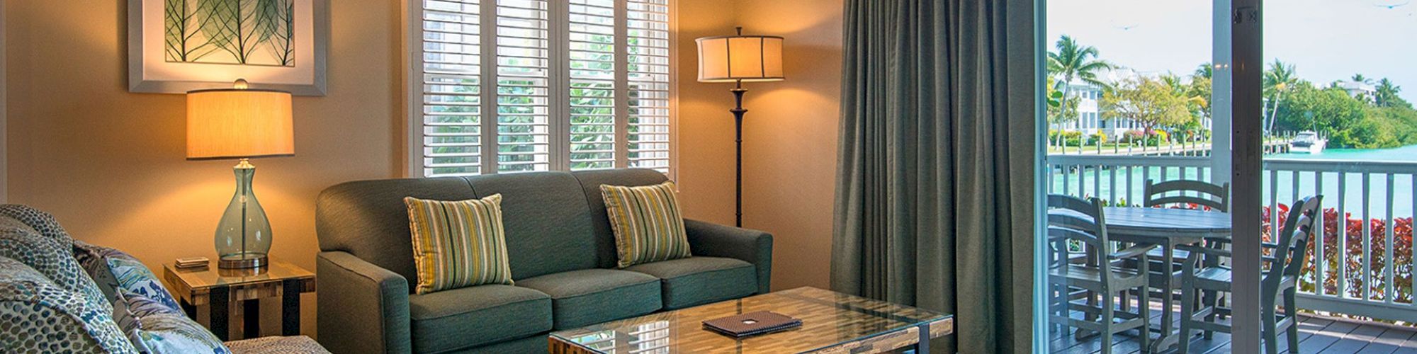 A well-lit living room with sofas, a coffee table, and lamps, beside a balcony overlooking a scenic outdoor view through sliding glass doors.