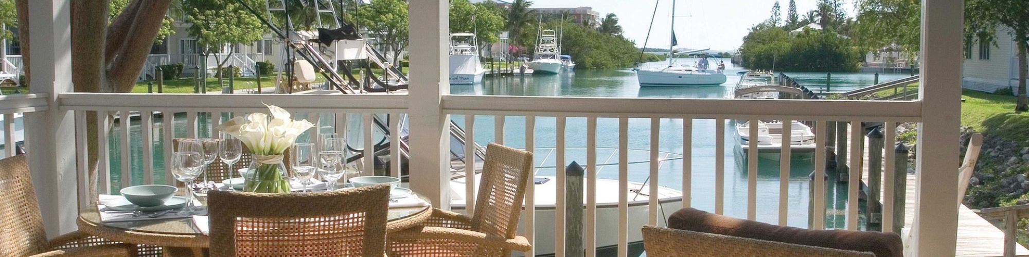 A serene outdoor patio with wicker chairs around a table overlooks a tranquil waterway with boats, framed by lush greenery in the background.