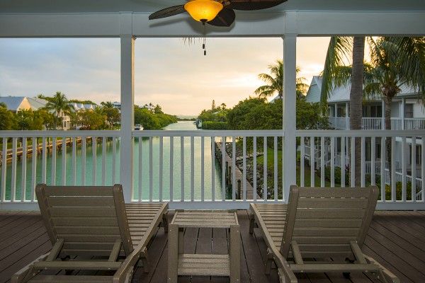 A serene balcony with two lounge chairs overlooks a river, surrounded by trees and buildings, under a soft evening sky.