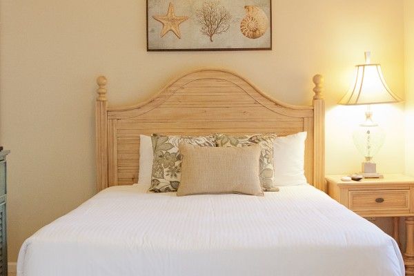 A cozy bedroom with a wooden headboard, three pillows on a white bed, a bedside lamp, and a wall art piece with sea-themed decor.