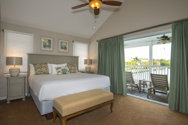 A cozy bedroom with a large bed, two side tables with lamps, a bench, a ceiling fan, and an open curtain leading to a balcony with chairs and a table.