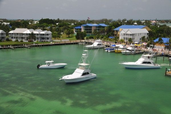 The image shows boats docked in a clear greenish marina, with residential houses and greenery in the background, under a cloudy sky.