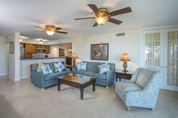 The image features a living room with two ceiling fans, a couch, an armchair, a coffee table, and lamps. The kitchen is visible in the background.